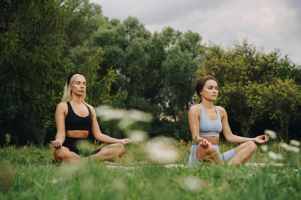Two women practicing yoga meditation in a serene outdoor park setting.